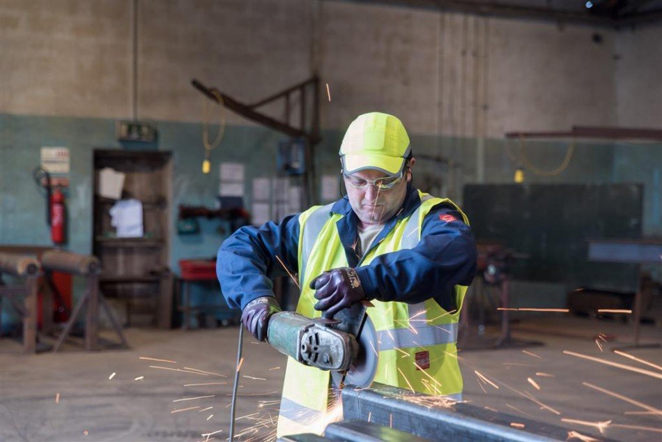 Worker in high-visibility clothing and a hard hat grinding a steel section inside the workshop