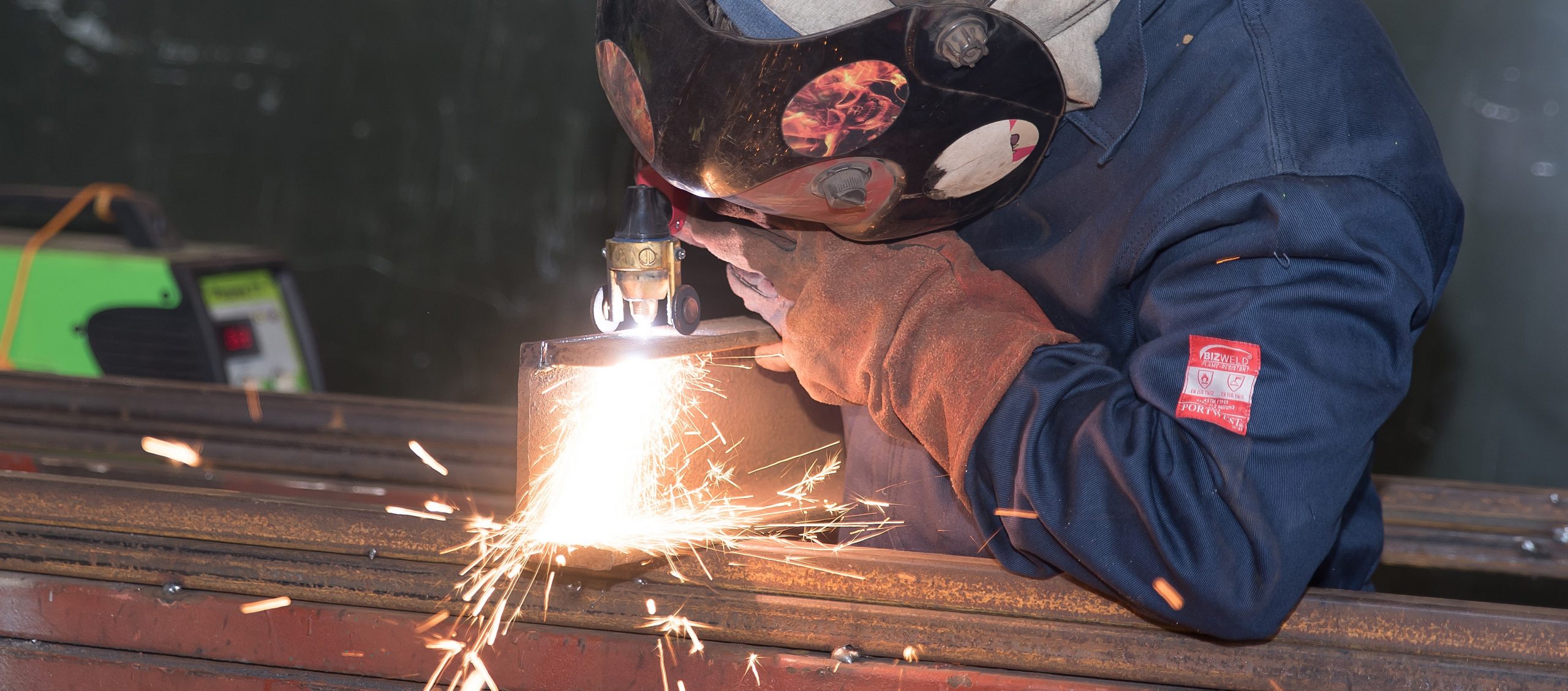 Welder in navy overalls with the mask down, sparks coming off a steel section on the bench